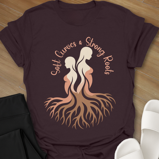 Brown t-shirt with Illustration of two curvy women with intertwined roots and the words “Soft Curves Strong Roots” in peach tones on a black background